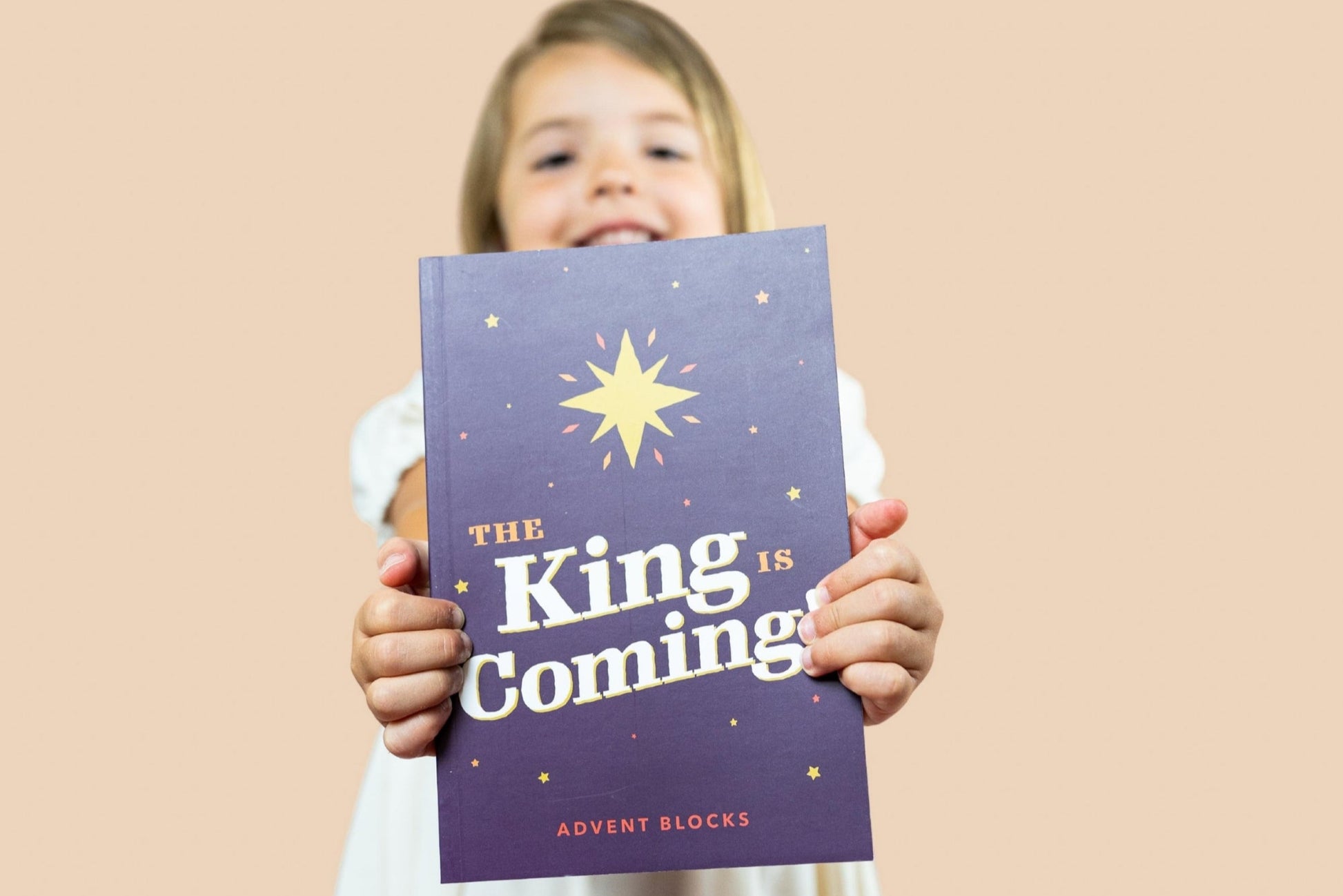Child holding a book titled 'The King is Coming' against a beige background